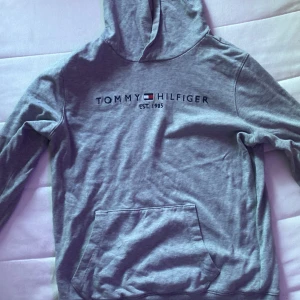Grå hoodie Tommy Hilfiger - Snygg grå hoodie från Tommy Hilfiger med broderad logga och text på bröstet. Klassisk känguruficka framtill och huva. Perfekt för en chill och avslappnad stil. Mjuk bomullskänsla och långärmad modell.