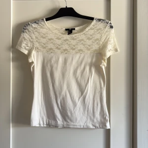 Vit spetsblus från H&M, storlek S - Supersöt vit blus från H&M i storlek S. Toppen har korta ärmar och en snygg spetsdetalj över axlar och bröst, medan resten är i mjuk bomull. Perfekt för dig som gillar romantiska och stilrena plagg med det lilla extra.