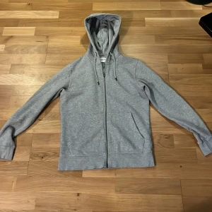Grå hoodie från Jack & Jones - Säljer en stilren grå hoodie från Jack & Jones med dragkedja framtill och huva med snörning. Tröjan har långa ärmar och två fickor på sidorna. Perfekt för chill dagar eller när du vill ha en enkel look. Materialet är mjuk bomull som känns skönt mot huden.