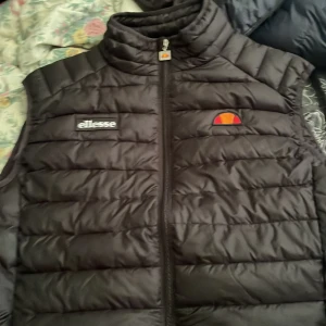 Svart dunväst från Ellesse, M - Snygg svart dunväst från Ellesse med broderad logga på bröstet och coolt märke på dragkedjan. Västen har quiltad design och hög krage, perfekt för lager-på-lager. Materialet är polyester och den stängs med dragkedja framtill. köpt för 3-4 år sen ny pris 1095 andvänt några gånger för länge sen 