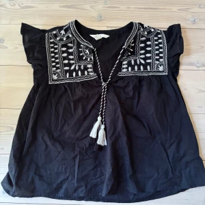 Svart blus med broderi från H&M - Svart blus från H&M med vit broderad detalj på bröstet och axlarna. Super fin och använd 2 gånger, perfekt till stranden 