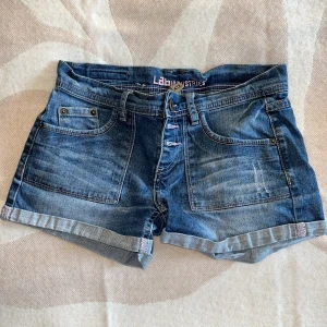 Blå jeansshorts med broderad ficka - Snygga blå jeansshorts från Lab Industries med uppvikta ben och coola broderade detaljer på bakfickorna. Klassisk femficksmodell med knappar framtill och slitningar för en avslappnad vibe. Perfekta för varma dagar och chill häng.