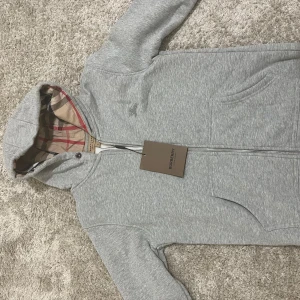 Grå hoodiejacka från Burberry - Grå hoodiejacka från Burberry med klassisk rutig insida i huvan. Jackan har dragkedja framtill, två fickor och broderad logga på bröstet. Mjuk bomullskänsla och snygg passform, perfekt för chill dagar. Ikonisk Burberry-tag i nacken.