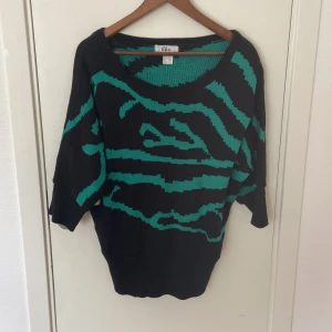 Svart-grön stickad tröja från b.p.c. - Cool stickad tröja från b.p.c. i svart med grönt zebramönster. Tröjan har bred halsringning och lite loose passform med trekvartsärmar. Perfekt för dig som gillar att sticka ut med mönster och färg. Snygg att matcha med jeans eller kjol.
