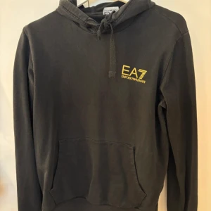 Svart EA7 hoodie med guld logga - Svart hoodie från EA7 Emporio Armani med gul logga på bröstet och längs ena ärmen. Tröjan har huva med snörning och en stor magficka framtill. Mjuk bomullskänsla och långärmad modell, perfekt för chill dagar eller streetwear. Finns ingen lapp med storlek men skulle gissa S-M