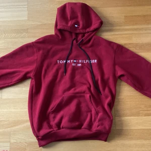 Mörkröd hoodie Tommy Hilfiger L - Säljer en mörkröd hoodie från Tommy Hilfiger i storlek L. Tröjan har broderad logga på bröstet, svart snörning i huvan och en stor magficka. Materialet är mjuk bomull och polyester, perfekt för chill dagar. Klassisk Hilfiger-flagga på huvan för extra stil.