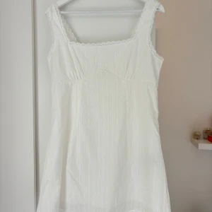 Vit kort klänning med spetskant - Supersöt vit kort klänning med tunna vertikala ränder och spetskant runt halsringning och axelband. Klänningen har en fyrkantig urringning och är ärmlös, med en lätt figurnära passform. Perfekt för varma dagar och sommarhäng.