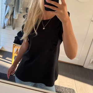 Svart blus med volangdetaljer - Säljer en svart blus med snygga volangdetaljer längs axlarna och ner mot bröstet. Blusen har rund hals och är kortärmad, vilket gör den enkel att matcha med jeans eller kjol. Materialet känns mjukt och har ett fint fall.