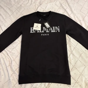 Balmain Paris sweatshirt - Svart sweatshirt från Balmain Paris med stor vit logotyp framtill. Tröjan har rund hals, långa ärmar och är tillverkad i mjuk bomull. Perfekt för dig som gillar stilrena designer och vill ha ett statement-plagg i garderoben.