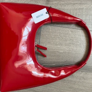 Röd axelväska från Bik Bok - Snygg röd axelväska från Bik Bok med glansig finish och stilren, trekantig form. Väskan har en matchande röd dragkedja och ett smidigt handtag som gör den enkel att bära. Perfekt för att addera färg till din outfit.