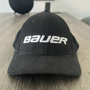 Svart Bauer keps med broderad logga - Snygg svart keps från Bauer med böjd skärm och vit broderad logga framtill. Kepsen har klassisk form och är tillverkad i bomull, med extra Bauer-detaljer på insidan och baksidan. Perfekt för dig som gillar sportig stil och vill ha något enkelt men stilrent.