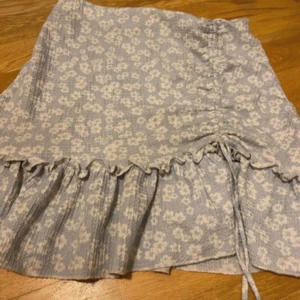 Blommig ljusblå kortkjol med volang - Supersöt ljusblå kortkjol med vita blommor och volangkant längst ner. Kjolen har snörning på sidan som ger en snygg detalj och extra form. Perfekt för dig som gillar romantisk och feminin stil. Mjuk och skön bomullskänsla. Skriv om ni har frågor!