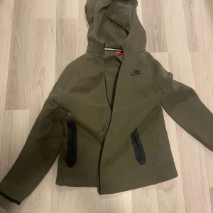 Olivgrön Nike vindjacka med huva - Snygg olivgrön vindjacka från Nike med svart logga på bröstet. Jackan har huva, två svarta dragkedjefickor och stilren design. Perfekt för dig som gillar sportig och clean look. Långärmad och med sköna muddar vid ärmslut.