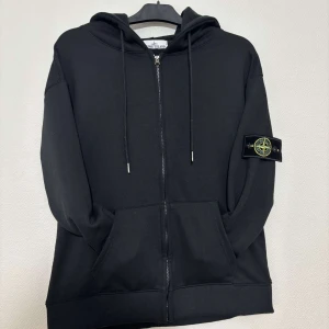 Svart hoodie från Stone Island - Svart hoodie från Stone Island med dragkedja och klassisk logotyp-patch på vänster ärm. Tröjan har huva med snörning och två stora fickor framtill. Tillverkad i mjuk bomull, perfekt för chill dagar. Snygg och enkel design som funkar till det mesta.