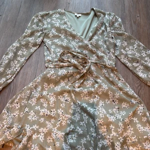 Blommig omlottklänning från Bubbleroom - Supersöt ljusgrön omlottklänning från Bubbleroom med vita blommor, lång ärm och knytband i midjan. Klänningen har v-ringad hals och volangdetaljer längst ner. Perfekt för vår och sommar när du vill känna dig extra fin.