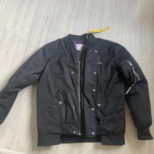 Svart bomberjacka Moncler med röd print - Svart bomberjacka från Moncler med fet röd graffiti-inspirerad print på ryggen och coola detaljer som dragkedja på ärmen och ribbade muddar. Jackan har fickor med tryckknappar och en schysst lila insida. Perfekt för dig som vill sticka ut.