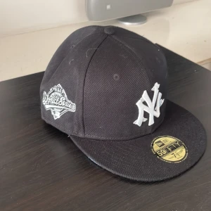 Svart New Era Yankees keps 59FIFTY - Snygg svart New Era 59FIFTY keps med New York Yankees broderat i vitt framtill och World Series patch på sidan. Platt skärm med ljusblå undersida och klassisk passform. Perfekt för dig som gillar streetstyle och baseball. Kepsen är oanvänd. 