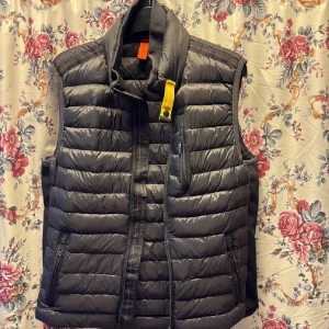 Mörkgrå dunväst Parajumpers  - Snygg mörkgrå dunväst från Parajumpers med quiltad design och cool gul detalj vid kragen. Västen har dragkedja fram, flera fickor med dragkedja och en hög krage. Perfekt för lager-på-lager och chill stil under höst och vinter.