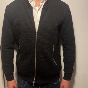 Svart zip up kofta från J.Lindeberg - OSÄKER PÅ PRIS SÅ KOM MED FÖRSLAG!!Säljer en svart zip up kofta från J.Lindeberg med snyggt strukturerat tyg och silvriga dragkedjor. Koftan har ribbade muddar och klassisk passform. Perfekt för dig som vill ha en stilren och modern look. 