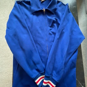 Blå bomberjacka med brodyr - Snygg blå bomberjacka med dragkedja framtill och broderad text 'DECORAH' i rött och vitt på ryggen. Jackan har klassisk krage och muddar med röd, vit och blå rand. Perfekt för dig som vill sticka ut med sportig retrostil.