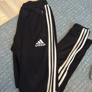 Svarta Adidas träningsbyxor YXL - Svarta Adidas träningsbyxor med vita ränder längs benen och klassisk logga på låret. Byxorna har elastisk midja, dragkedja vid fickan och är gjorda i lätt polyester med Climacool-teknologi som andas. Perfekta för sport eller chill.