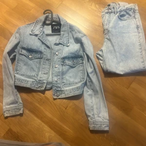 Ljusblå croppad jeansjacka från Zara - Säljer en ljusblå croppad jeansjacka från Zara med klassisk krage, två stora bröstfickor med knappar och snygga detaljer vid midjan. Jackan har långa ärmar och silverfärgade knappar framtill. Perfekt för dig som gillar en chill och trendig look.