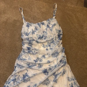 Vit och blå blommig långklänning - Säljer en superfin klänning med smala axelband och blommigt mönster i blått på vit botten. Klänningen har en rynkad och figurnära passform som ger en snygg siluett. Stretchig så passar XS,S och M. Aldrig använd 