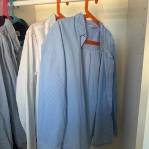 Blå och vit skjorta från C, 146/ 11 years old.  - Säljer två skjortor från C i storlek for 11 år pojkar. Ena är vit med liten broderad logga på bröstet, den andra är ljusblå med smala vita ränder. Båda har klassisk krage, knappar framtill och långa ärmar. Perfekt för dig som gillar stilrena och enkla plagg.     50kr för båda. 30kr för en 