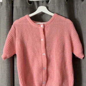 Rosa stickad kofta med kort ärm - Superfin rosa stickad kofta med korta ärmar och rund hals. Koftan har en ribbad struktur och knäpps framtill med vita knappar. 