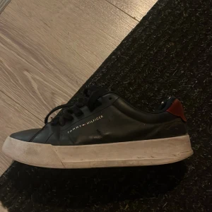 Svarta sneakers Tommy Hilfiger - Snygga svarta sneakers från Tommy Hilfiger med vit platt sula och röd detalj vid hälen. Skon har snörning och är tillverkad i skinn, perfekt för dig som gillar stilrena och klassiska sneakers med en twist.