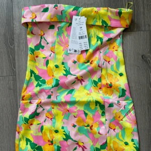 Blommig axelbandslös kortklänning - Färgstark axelbandslös kortklänning från Gina med blommigt mönster i rosa, gul, grön och inslag av orange. Klänningen har en snygg omlottkant upptill och är gjord i glansig satin som känns lyxig mot huden. Perfekt för sommarens alla tillfällen!