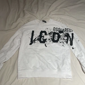 Vit Dsquared2 ICON sweatshirt - Säljer en vit sweatshirt från Dsquared2 med stor svart ICON-print och coolt splattermönster över bröstet. Tröjan har rund hals, långa ärmar och är gjord i mjuk bomull. Perfekt för dig som gillar streetstyle och vill sticka ut med ett statement-plagg.
