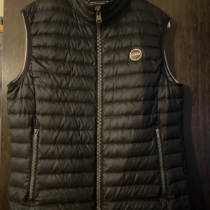 Colmar väst - Snygg svart dunväst från Colmar Originals med quiltad design och två praktiska fickor med dragkedja. Västen har hög krage och logotyp på bröstet. Perfekt att slänga över en hoodie för extra värme och stil.