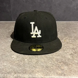 Svart LA-keps från New Era 59FIFTY - Säljer en svart LA Dodgers keps från New Era, modell 59FIFTY. Klassisk flat brim och broderad LA-logga i vitt framtill. MLB-logga baktill och New Era-logga på sidan. Snygg streetstil och passar dig som gillar sport och urban vibe.