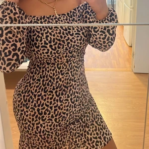 Leopardmönstrad offshoulder klänning - Säljer en kort, leopardmönstrad klänning med offshoulder och puffiga långärmar. Klänningen har en figurnära passform och är perfekt för dig som vill sticka ut. Färgerna går i beige och svart och materialet känns mjukt och följsamt.