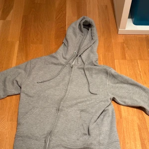 Grå hoodie från Pull&Bear med dragkedja - Säljer en enkel och snygg grå hoodie från Pull&Bear i storlek XS. Tröjan har huva med snörning, praktiska fickor framtill och hel dragkedja. Perfekt för chill dagar eller när du vill ha något bekvämt och stilrent. Materialet är mjuk bomullsmix.
