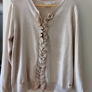 Beige kofta med volang från Holly & Whyte - Supersöt beige kofta från Holly & Whyte med volangdetalj längs knäppningen framtill. Koftan har långärmade och ribbade muddar vid ärmslut och nederkant. Snygg metallbricka med logga vid nederkanten.