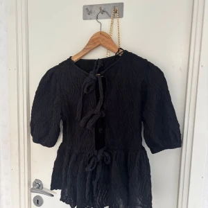 Svart blus Gina tricot  - Svart blus med tre knyt framtill och puffiga halvlånga ärmar. Tyget har en snygg strukturerad yta och blusen har en lätt volang nertill. Perfekt för dig som gillar unika detaljer och en trendig look.
