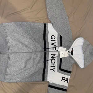 Gicenchy Paris kofta - Snygg hoodie från Givenchy i grått med vita och svarta paneler och broderad logga över bröstet. Tröjan har huva med snörning, dragkedja framtill och stora fickor. Perfekt för dig som gillar streetwear och exklusiva märken. Helt oanvänd och ny kommer med taggarna! Priset kan förhandlas vi snabbaffär