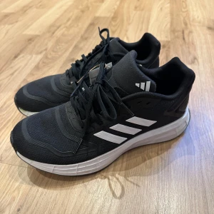 Svarta helt nya Adidas sneakers med vita ränder - Säljer ett par helt nya svarta Adidas sneakers med klassiska vita ränder på sidorna och vit sula. Skorna har snörning och är tillverkade i syntetmaterial med mesh-överdel för bra ventilation. Perfekta för dig som gillar sportig stil och vill ha bekväma skor till träning eller häng. Min son skulle använda skorna för första gången idag men då han har växt otroligt mycket i sommar så var skorna för små, använt bara tofflor under sommaren. Så använda 1 timme idag. 