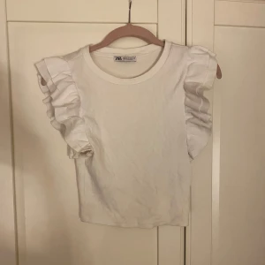 Vit volangärmad topp från Zara - Supersöt vit topp från Zara i ribbad bomull med volangdetaljer på axlarna. Modellen är croppad och har rund hals. Perfekt för dig som gillar en clean look med lite extra edge på ärmarna. Säljer då den inte kommer till användning! 💘💘