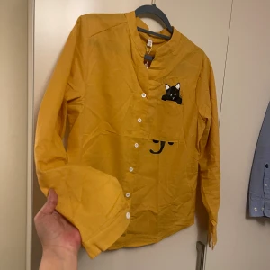 Gul henley med kattmotiv - Säljer en cool gul henley-skjorta med lång ärm och vita knappar. Skjortan har ett unikt tryck med en svart katt som kikar ut ur bröstfickan. Materialet är mjuk bomull och modellen har en avslappnad passform. Perfekt för dig som gillar roliga detaljer!helt ny Storlek L40