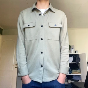 Olivgrön overshirt från Selected Homme - Snygg olivgrön overshirt från Selected Homme med svarta knappar och två bröstfickor med lock. Skjortan har klassisk krage och lång ärm, perfekt att bära över en t-shirt. Materialet är mjukt och känns som bomull. Enkel och stilren design som funkar till jeans. Modellen på bilden är 184cm, 65kg. Använd fåtal gånger men helt helt som nyskick! Nypris 800kr. Hör av dig vid minsta fundering!🔥