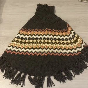 Mönstrad stickad poncho från H&M - Säljer en sjukt mysig stickad poncho från H&M med höga polokrage och fransar längs kanten. Färgerna är mörkgrå, vit, rosa och gul med ett snyggt zigzag-mönster och inslag av glittrigt garn. Perfekt för kalla dagar när du vill vara både varm och stilren.