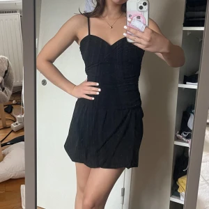 Svart kort klänning med smala axelband - Supersnygg svart klänning med smala axelband och v-ringad överdel. Sitter figurnära upptill med draperade detaljer och har en utsvängd kjol. 