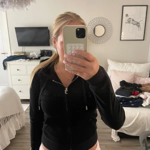 Svart Juicy Couture tröja - Svart hoodie från Juicy Couture i mjuk velour med huva och snörning. Dragkedja framtill med ikonisk J-detalj. Klassisk, slim passform och ribbade muddar. Perfekt för chill dagar och snygg streetstyle.