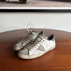 Philippe Model vita sneakers med gröna detaljer - Snygga sneakers från Philippe Model i vitt och grått med gröna detaljer på hälen. Skorna har rund tå, snörning och platt sula. Materialet är en mix av skinn och mocka, vilket ger en cool och urban vibe. Perfekta för dig som gillar streetstyle och vill sticka ut. 