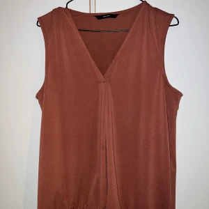 Brun ärmlös blus från Vero Moda - Snygg brun blus från Vero Moda med v-ringad hals och ett lätt draperat fall framtill. Modellen är ärmlös och har en mjuk, slät yta i polyester med lite stretch för extra komfort. Perfekt att styla med jeans eller kjol för en chill look.