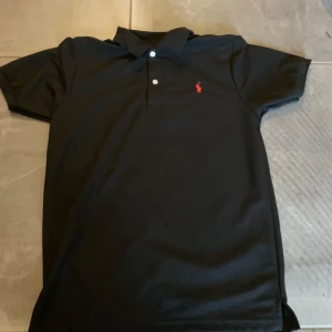 Polo Ralph lauren piké  - Svart pikétröja från Ralph Lauren med klassisk krage och två vita knappar framtill. Tröjan har korta ärmar och är gjord i mjuk bomull. På bröstet finns den ikoniska röda broderade polospelaren. Perfekt för en clean och stilren look. Storleken är M och neck tagen är bortklippt därav det billiga priset 