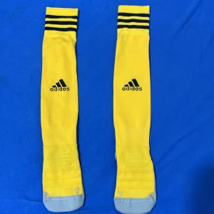 Gula fotbollsstrumpor från Adidas - Säljer ett par långa gula fotbollsstrumpor från Adidas med svarta ränder upptill och grå förstärkning vid tår och häl. Klassisk Adidas-logga på sidan. Perfekta för match eller träning, sitter skönt och har ribbad struktur vid vristen.